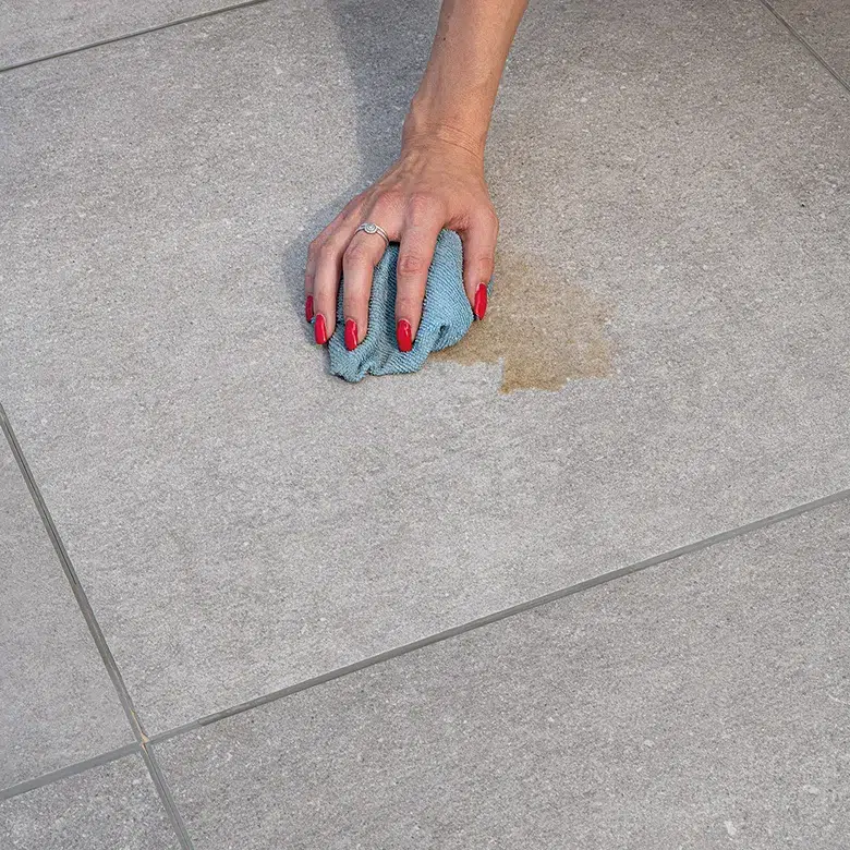 Hand with red nails wiping a brown stain from light grey porcelain tiles with a blue cloth, demonstrating easy cleaning of interlocking floor.
